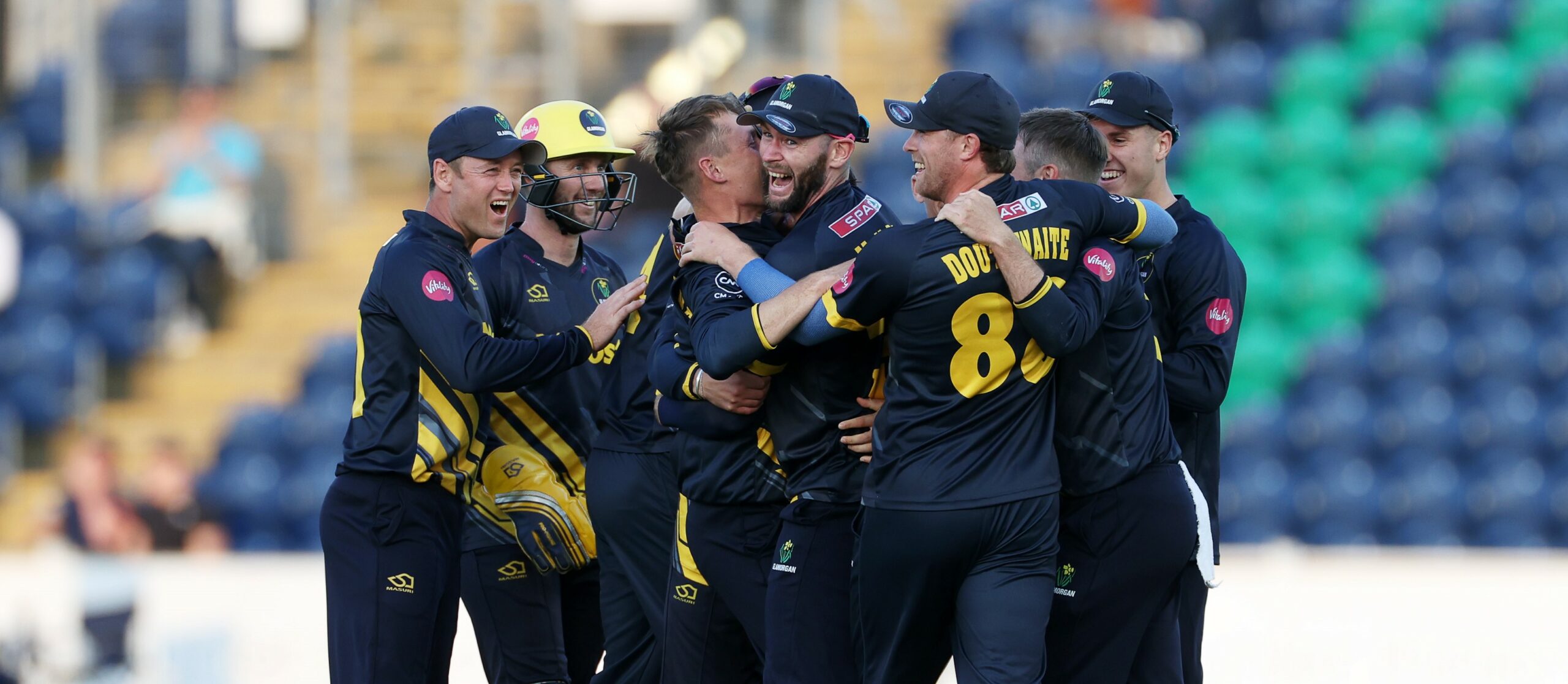 A cricket team celebrate and hug each other on the pitch