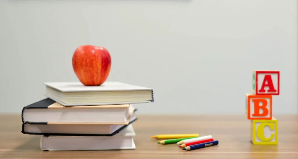 A desk in a classroom with books, pencils, ABC blocks and an apple on it.