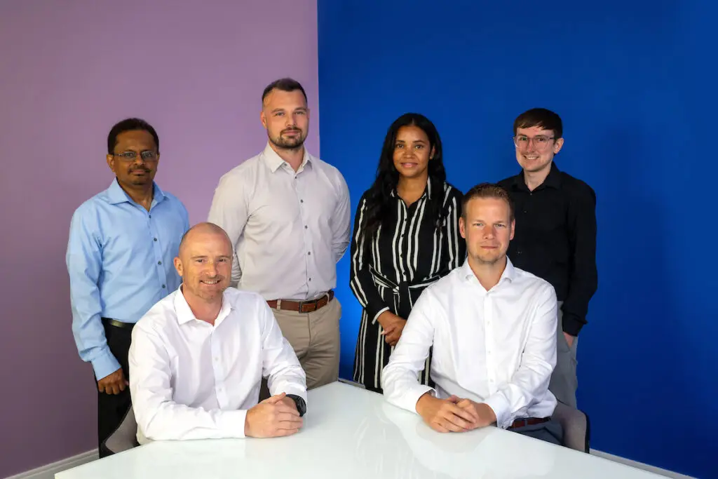 Four employees stand in front of purple and blue walls. There are two senior male employees sat in front of them at a white table