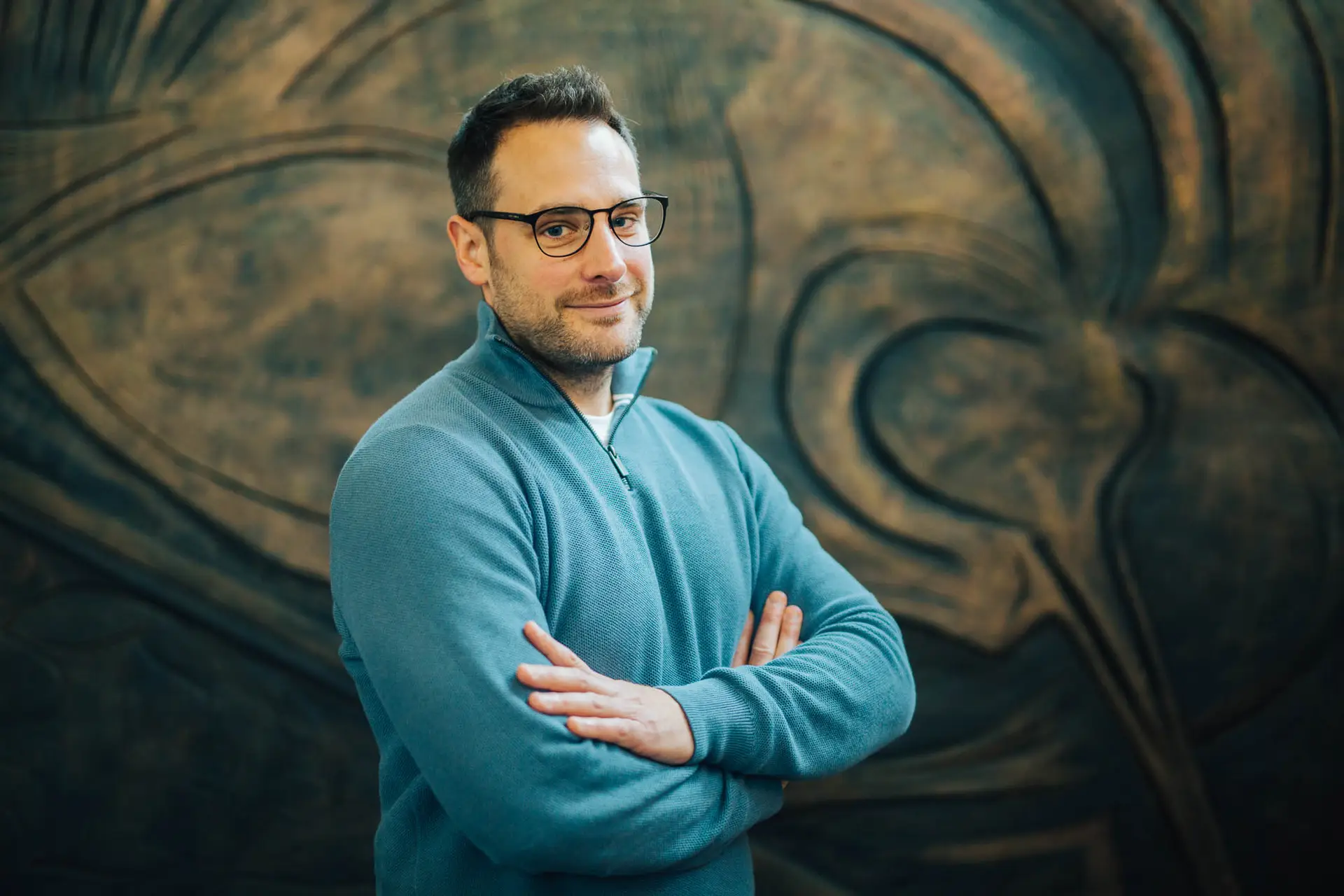 A man stands with his arms folded. He is wearing glasses and a blue fleece.