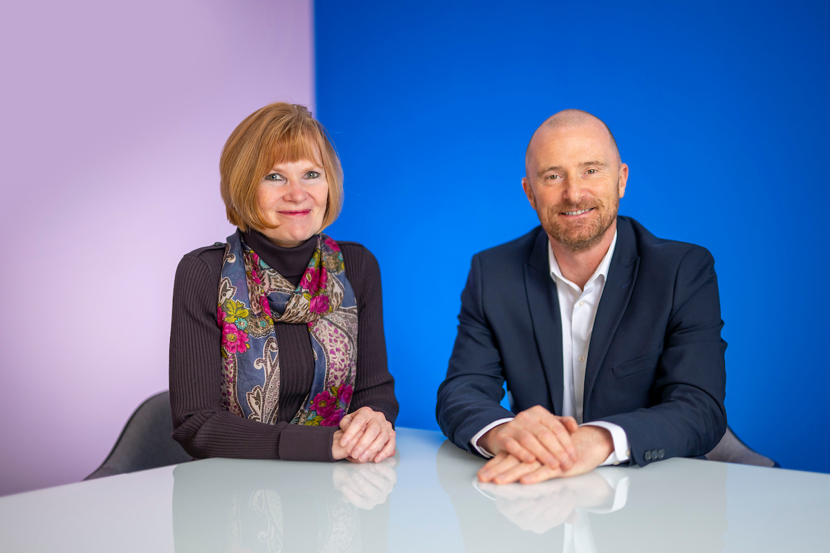 A woman in a dark top and colourful scarf sits next to a man in a suit without a tie.