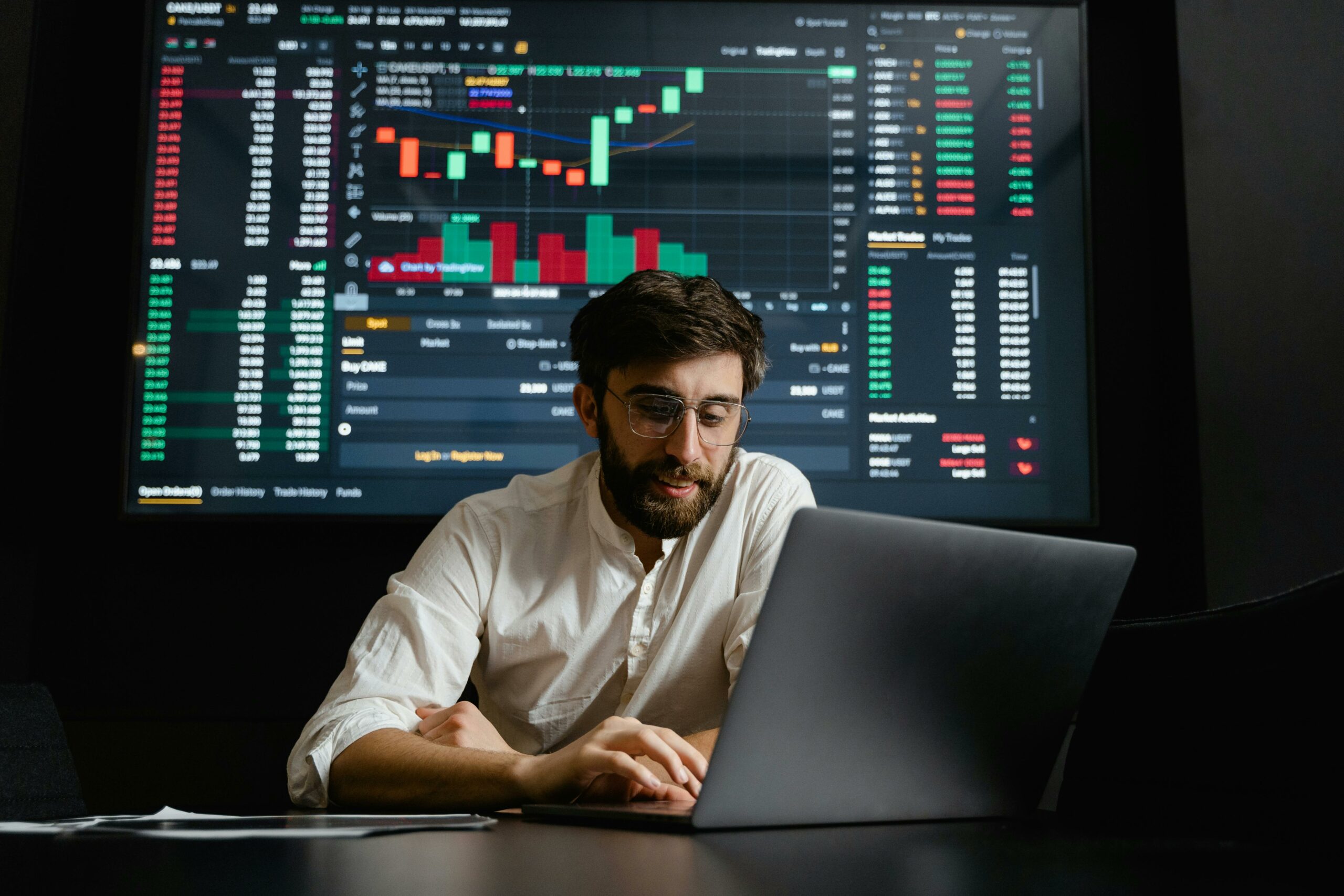 A brunette man wearing a white shirt looking at a laptop. A board behind him shows various graphs and numerical figures.