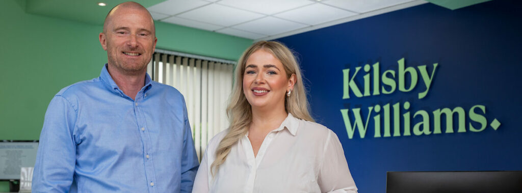 A woman in a white shirt and black skirt leans on a desk. She is stood next to a man in a blue shirt and pale trousers.